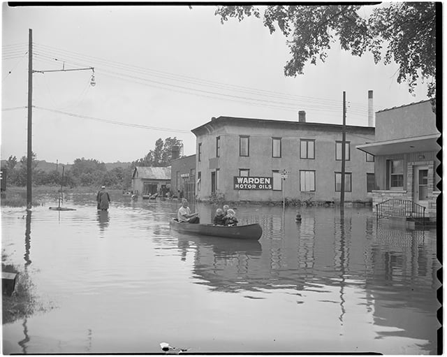Black-and-white photograph of a man and three young children in a canoe, paddling through glassy water in a flooded street. Water is up to the first few feet of the two utility poles and four buildings in the photo; two men in the background wade through water up to their knees. 